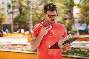 Poster - Young hispanic man talking on the smartphone reading document at park