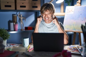 Canvas Print - Middle age man sitting at art studio with laptop at night covering ears with fingers with annoyed expression for the noise of loud music. deaf concept.