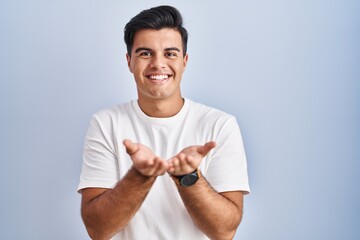 Sticker - Hispanic man standing over blue background smiling with hands palms together receiving or giving gesture. hold and protection