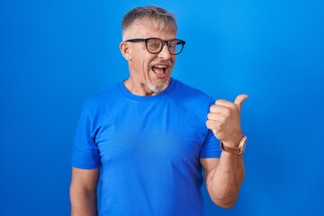 Wall Mural - Hispanic man with grey hair standing over blue background smiling with happy face looking and pointing to the side with thumb up.