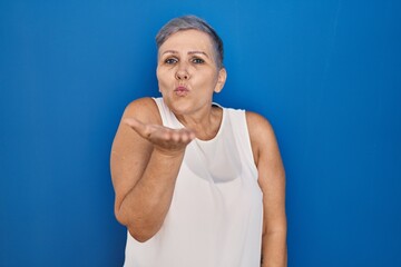 Wall Mural - Middle age caucasian woman standing over blue background looking at the camera blowing a kiss with hand on air being lovely and sexy. love expression.