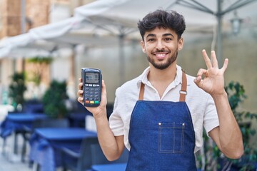 Poster - Arab man with beard wearing waiter apron at restaurant terrace holding dataphone doing ok sign with fingers, smiling friendly gesturing excellent symbol