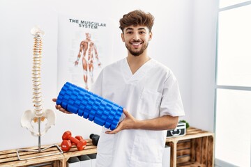 Poster - Young arab man wearing physiotherapist uniform holding foam roller at clinic