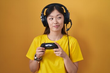 Wall Mural - Chinese young woman playing video game holding controller smiling looking to the side and staring away thinking.