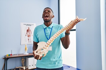 Poster - African american man holding anatomical model of spinal column angry and mad screaming frustrated and furious, shouting with anger looking up.