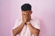 Young hispanic man standing over pink background with sad expression covering face with hands while crying. depression concept.