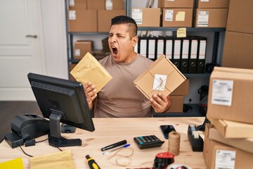 Canvas Print - Hispanic young man working at small business ecommerce holding packages angry and mad screaming frustrated and furious, shouting with anger. rage and aggressive concept.