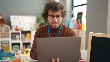 Poster - Young hispanic man preschool teacher using laptop standing at kindergarten