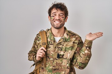 Canvas Print - Hispanic young man wearing camouflage army uniform smiling cheerful presenting and pointing with palm of hand looking at the camera.