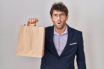 Sticker - Hispanic business man holding delivery bag scared and amazed with open mouth for surprise, disbelief face