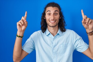 Poster - Young hispanic man standing over blue background smiling amazed and surprised and pointing up with fingers and raised arms.