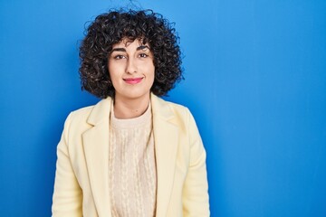 Poster - Young brunette woman with curly hair standing over blue background relaxed with serious expression on face. simple and natural looking at the camera.