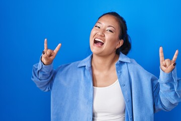 Wall Mural - Asian young woman standing over blue background shouting with crazy expression doing rock symbol with hands up. music star. heavy music concept.