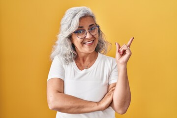 Wall Mural - Middle age woman with grey hair standing over yellow background with a big smile on face, pointing with hand and finger to the side looking at the camera.