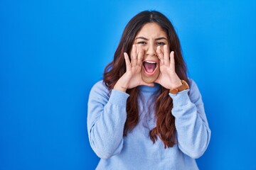 Canvas Print - Hispanic young woman standing over blue background shouting angry out loud with hands over mouth