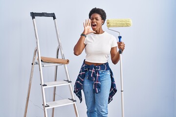 Poster - African american woman holding roller painter shouting and screaming loud to side with hand on mouth. communication concept.