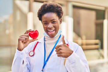 Poster - African american woman wearing doctor uniform holding heart smiling happy and positive, thumb up doing excellent and approval sign