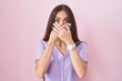 Young hispanic woman with long hair standing over pink background shocked covering mouth with hands for mistake. secret concept.