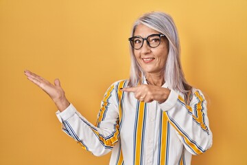 Canvas Print - Middle age woman with grey hair standing over yellow background wearing glasses amazed and smiling to the camera while presenting with hand and pointing with finger.