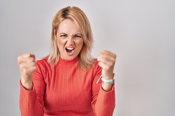 Wall Mural - Blonde woman standing over isolated background angry and mad raising fists frustrated and furious while shouting with anger. rage and aggressive concept.