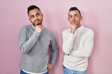 Poster - Homosexual couple standing over pink background with hand on chin thinking about question, pensive expression. smiling with thoughtful face. doubt concept.