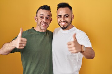 Wall Mural - Homosexual couple standing over yellow background approving doing positive gesture with hand, thumbs up smiling and happy for success. winner gesture.