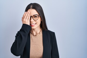 Canvas Print - Young brunette woman standing over blue background covering one eye with hand, confident smile on face and surprise emotion.