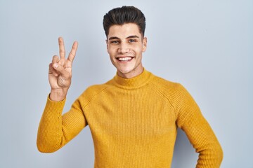 Canvas Print - Young hispanic man standing over blue background smiling looking to the camera showing fingers doing victory sign. number two.