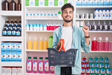 Sticker - Young hispanic man with tattoos shopping at pharmacy drugstore holding basket smiling with an idea or question pointing finger with happy face, number one