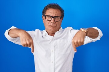 Wall Mural - Middle age hispanic man standing over blue background pointing down looking sad and upset, indicating direction with fingers, unhappy and depressed.