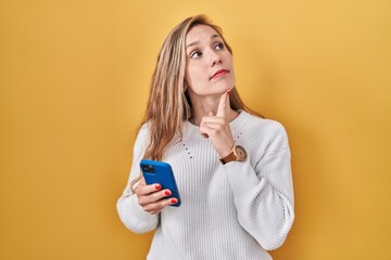 Wall Mural - Young blonde woman using smartphone typing message with hand on chin thinking about question, pensive expression. smiling with thoughtful face. doubt concept.