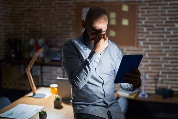 Wall Mural - Young hispanic man with beard and tattoos working at the office at night tired rubbing nose and eyes feeling fatigue and headache. stress and frustration concept.