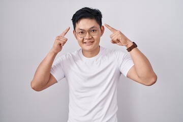 Wall Mural - Young asian man standing over white background smiling pointing to head with both hands finger, great idea or thought, good memory