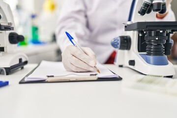 Wall Mural - Middle age hispanic woman wearing scientist uniform using microscope write on document at laboratory