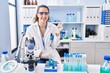 © Krakenimages.com - Young hispanic woman working at scientist laboratory with a big smile on face, pointing with hand and finger to the side looking at the camera.
