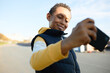 © Anatoliy Karlyuk - Selective focus of handsome smiling black boy blogger of 13-years-old in hoodie and vest taking selfie on his smartphone or recording stories, making content against urban landscape background