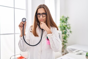 Poster - Young woman wearing doctor uniform holding stethoscope serious face thinking about question with hand on chin, thoughtful about confusing idea