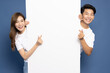 © comzeal - Young Asian couple is standing behind the white blank banner or empty copy space advertisement board and showing mini heart sign isolated on blue background, Looking at camera