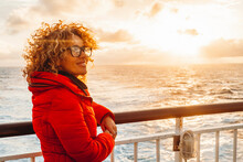 Portrait Of A Cheerful Young Adult Woman Enjoying Sunset Outside Traveling The Ocean On A Ferry Cruise Ship Boat Alone. Traveler People Lifestyle, Red Jacket. Orange Warm Sky Background. Transport