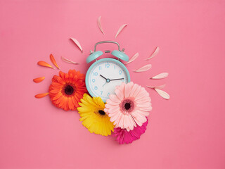 Spring time concept. Creative flat design with turquoise alarm clock on pink background with vibrant colorful flowers around it and some petals detached on top. Flat design, top view, copy space.