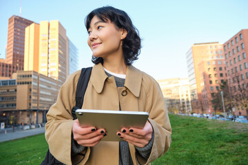 Wall Mural - Happy young brunette girl, asian woman walks around city with tablet, goes to university with her digital gadget and backpack