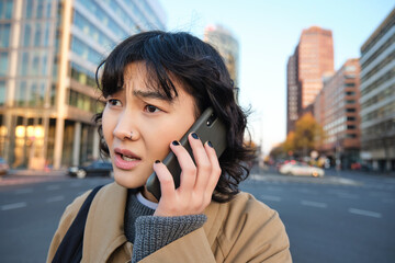 Wall Mural - Close up portrait of concerned asian woman, talks on mobile phone and hears bad news, looks worried, feels frustrated by telephone conversation, stands on street