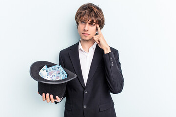 Young magician man holding a hat with banknotes isolated on blue background pointing temple with finger, thinking, focused on a task.