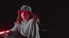 Young Female Dancer Dancing Outdoors At Night While Holding Pink Neon Lamp. Red-haired Girl In Gray Hoodie And Camouflage Hat Making Freestyle Dance Moves. Dancing, Culture, Youth Concept