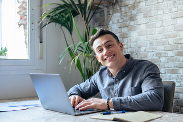 Wall Mural - Good-looking millennial office employee in glasses sitting at desk in front of laptop smiling looking at camera. Successful worker, career advance and opportunity, owner of prosperous business concept