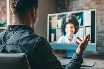 View over shoulder employer listen applicant at job interview online use cam and pc. Young caucasian man with tattoos at home working and talking online with African woman colleague in videocall.