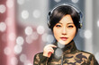 Young Asian woman in a camouflage suit listens to music with headphones on her ears, light reflections in the picture, fictional person, made with generative AI