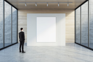 Wall Mural - Man in black suit back view looking at blank white poster with space for your logo or text on light partition on light wooden background in empty exhibition hall with transparent walls, mock up