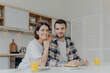© VK Studio - Positive brunette woman leans at shoulder of her husband, pose together at kitchen, enjoy delicious breakfast, have happy mood, look directly at camera, have good relationships. Family concept