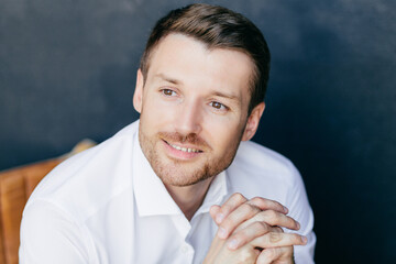 Wall Mural - Photo of cheerful pensive young male with bristle, dreams about something pleasant, wears white shirt, poses against dark wall. Successful prosperous male executive worker contemplates indoor